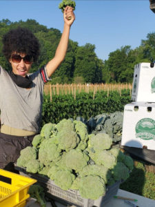 woman holding broccoli in the air and standing in front of a pile of broccoli heads