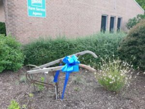 An antique farm tool in a garden with a blue ribbon attached.